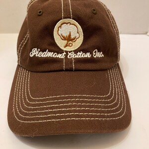 Puede incluir: Gorra de b&eacute;isbol marr&oacute;n con costuras blancas y el texto "Piedmont Cotton Inc.". La gorra presenta un parche circular con un dise&ntilde;o de planta de algod&oacute;n. La visera y la corona son marrones, con detalles de costura blancos.