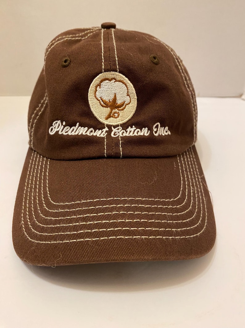 Puede incluir: Gorra de b&eacute;isbol marr&oacute;n con costuras blancas y el texto "Piedmont Cotton Inc.". La gorra presenta un parche circular con un dise&ntilde;o de planta de algod&oacute;n. La visera y la corona son marrones, con detalles de costura blancos.
