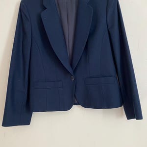 May include: A navy blue blazer with a single-button closure, two front pockets, and a notched lapel. The blazer has a tailored fit and a classic design, suitable for various occasions. The blazer is hanging on a black hanger.