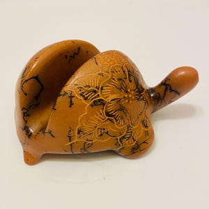May include: A handcrafted, turtle-shaped napkin holder. The holder is a warm brown color with black floral etchings. The turtle has a handle and a slot to hold napkins. The holder is made of a hard material.
