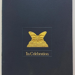 May include: A navy blue card with a gold embossed butterfly design and the words "In Celebration." The butterfly is centered within a rectangular frame. The card has a textured surface.