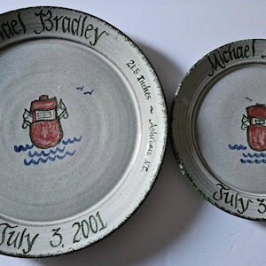 May include: Two hand-painted ceramic plates with a nautical theme. The plates are gray with a dark green rim and feature a red boat on blue waves. The larger plate includes the name "Michael Bradley", date, time, weight, and height.
