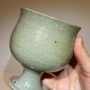 May include: A ceramic goblet with a speckled light green glaze and a textured, matte finish. The vessel features a wide, flared rim, a rounded bowl, and a sturdy pedestal base. The surface displays small dark brown flecks throughout the clay body.