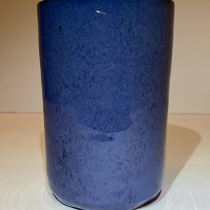 May include: A cylindrical ceramic tumbler featuring a deep blue glaze with a mottled, speckled texture throughout the surface. The vessel has a smooth, straight-sided profile with a rounded base and a consistent, uniform color distribution.