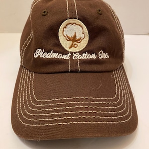 Puede incluir: Gorra de b&eacute;isbol marr&oacute;n con costuras blancas y visera curva. La gorra presenta un parche circular con un dise&ntilde;o de planta de algod&oacute;n y el texto "Piedmont Cotton Inc." en escritura blanca.