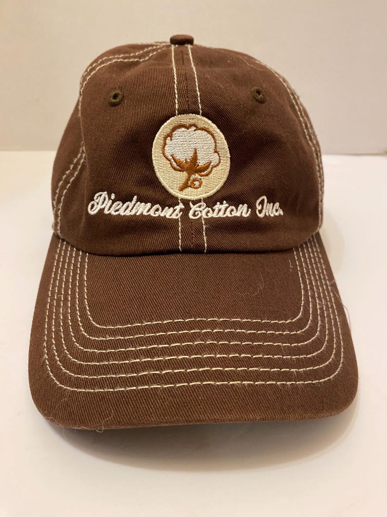 Puede incluir: Gorra de b&eacute;isbol marr&oacute;n con costuras blancas y visera curva. La gorra presenta un parche circular con un dise&ntilde;o de planta de algod&oacute;n y el texto "Piedmont Cotton Inc." en escritura blanca.