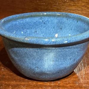 May include: A small, round ceramic bowl with a speckled blue glaze. The bowl features a smooth, curved interior and a slightly flared rim, showcasing a consistent mottled texture across its exterior surface and a glossy finish throughout the piece.