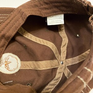 Puede incluir: Gorra de b&eacute;isbol marr&oacute;n con un emblema de planta de algod&oacute;n. El interior presenta tela marr&oacute;n con costuras beige y una etiqueta que dice "100% Cotton Made in Bangladesh".