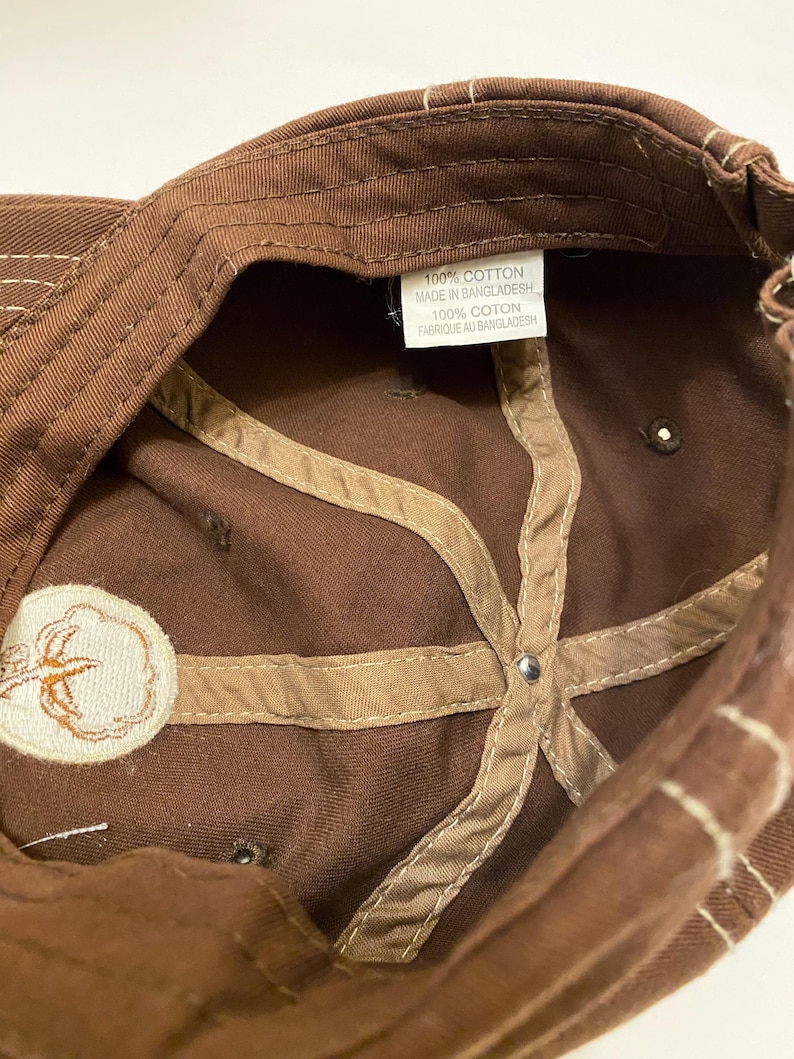 Puede incluir: Gorra de b&eacute;isbol marr&oacute;n con un emblema de planta de algod&oacute;n. El interior presenta tela marr&oacute;n con costuras beige y una etiqueta que dice "100% Cotton Made in Bangladesh".