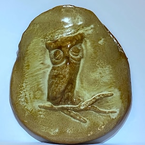 May include: A circular, tan-colored ceramic plaque featuring a raised, embossed relief of an owl perched on a branch. The surface has a glossy, glazed finish with variations in color intensity, highlighting the detailed texture of the owl.