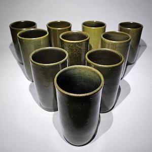 May include: A collection of ten handmade ceramic tumblers in varying shades of green. The cylindrical cups have a textured surface and a dark interior. The arrangement of the cups creates a triangular shape, showcasing their unique design and craftsmanship.