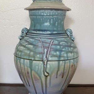 May include: A large, teal-colored ceramic vase with a lid. The vase features a textured design and decorative handles. The lid has a loop handle. The glaze has a dripped effect, with shades of blue, green, and brown. The vase sits on a dark wooden surface.