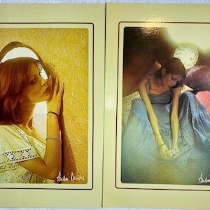 May include: Two vintage art prints featuring women. The first shows a woman in a light-colored top, touching a textured wall. The second depicts a woman in a blue dress, seated with flowers. Both prints have a warm color palette and are signed.