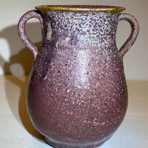 May include: A ceramic vase with two handles, featuring a textured, speckled surface in shades of deep purple and brown. The rim has a distinct yellow-gold accent, and the body has a rounded, bulbous shape with a slightly tapered neck and base.