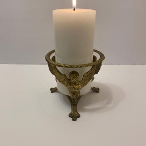 May include: A lit, white pillar candle in a decorative brass candle holder. The candle holder features an angel design and a twisted rope detail around the top. The candle is centered and burning brightly.