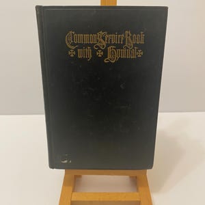 May include: A black book titled "Common Service Book with Hymnal" in gold lettering. The book is displayed on a small wooden easel. The background is a plain white surface.