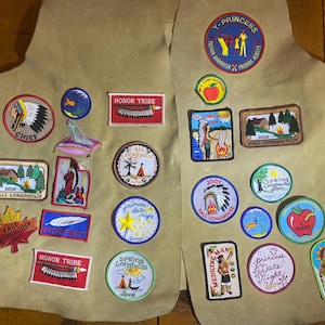 May include: Tan vest adorned with colorful patches. Patches feature text like "CHIEF", "HONOR TRIBE", "Y-PRINCESS", and "FALL LONGHOUSE". Designs include Native American imagery, a campfire, an apple, and a star. A unique item for collectors.