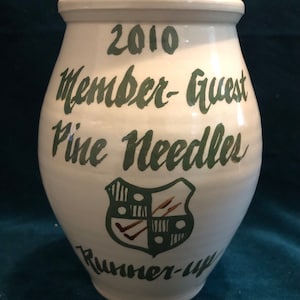 May include: White ceramic jar with green lettering that reads "2010 Member-Guest Pine Needles Runner-Up". The jar has a green and white shield with a brown design.