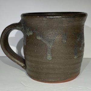 May include: A handmade ceramic mug with a dark brown glaze. The mug has a rounded body, a sturdy handle, and a reddish-brown base. The glaze has subtle variations in color, with hints of blue and green.