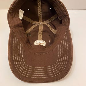 Puede incluir: Gorra de b&eacute;isbol marr&oacute;n con visera curva y correa ajustable. El interior tiene un forro de algod&oacute;n y un peque&ntilde;o emblema bordado. Las costuras blancas acent&uacute;an la visera y las costuras.