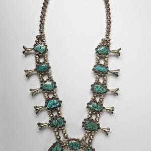May include: A silver squash blossom necklace with turquoise stones. The necklace features a beaded chain and a horseshoe-shaped pendant with turquoise stones. The turquoise stones are a vibrant blue-green color.