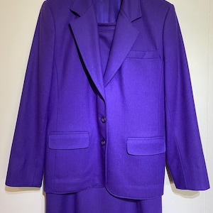 May include: A solid purple two-piece suit consisting of a long-sleeve blazer and a matching skirt. The blazer features a notched lapel, two front buttons, and two flap pockets. A Pendleton brand label is visible on the inside collar.