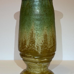 May include: A handcrafted ceramic vase with a green and brown glaze. The vase has a cylindrical shape with a flared base and a slightly wider top. The glaze has a textured appearance, with variations in color and tone.