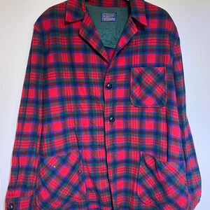 May include: A long-sleeve button-up shirt featuring a red, blue, and dark green plaid pattern. The shirt has a pointed collar, a single chest pocket on the left side, two lower front pockets, and dark buttons. A Pendleton brand label is visible.