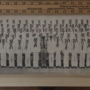 May include: Black and white photograph of a group of men in uniform, likely military personnel, standing in formation. They are wearing white shirts with black ties and hats. A flag with a number "2" on it is visible in the front right corner of the image. The text "UNIT NO. 2 BATTALION, U.S. NAVAL TRAINING STATION, GREAT LAKES, ILL." is printed below the image.