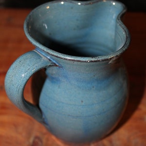 May include: A blue ceramic pitcher with a wide mouth and a handle. The pitcher has a speckled glaze and a slightly uneven surface.