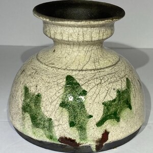 May include: A ceramic vase with a cream-colored body and a dark brown rim and base. The vase features green tree-like designs and a cracked glaze effect, adding a rustic charm to the piece.