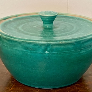 May include: A handcrafted, teal-colored ceramic bowl with a matching lid. The bowl has a rounded shape and a slightly textured surface. The lid features a small, rounded handle. The bowl is approximately 6 inches in diameter.