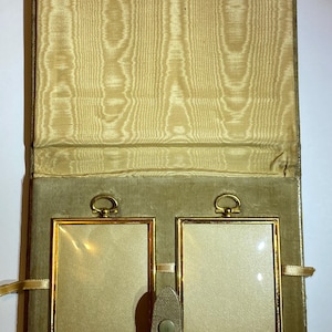 May include: A folding double picture frame case with a beige moiré fabric interior and a soft olive-green velvet lining. The case contains two rectangular gold-toned metal frames with top loops, secured by a central olive-green snap-button tab.