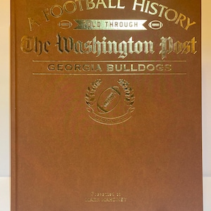 May include: A brown hardcover book titled A Football History Told Through The Washington Post Georgia Bulldogs with gold foil lettering and a central football emblem surrounded by a laurel wreath, featuring the text Presented to MARK MAHONEY.