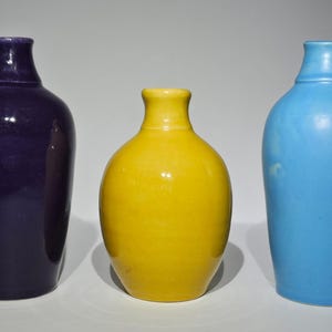 May include: Three ceramic vases in a row, each with a unique color. The vase on the left is a deep purple, the middle vase is yellow, and the vase on the right is light blue. All vases have a similar shape.