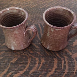 May include: Two brown ceramic mugs with a speckled glaze and a unique handle design.