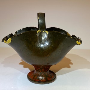 May include: A dark olive green ceramic basket with a central handle and a pedestal base. The rim features scalloped edges with visible gold-colored accents. The glaze has a mottled texture, and the base shows reddish-brown undertones.