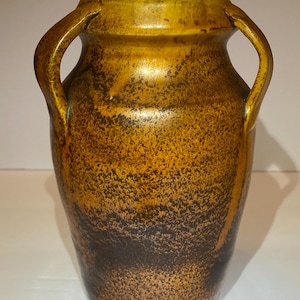 May include: A ceramic vase featuring two side handles and a wide opening. The exterior surface displays a mottled, textured finish with a gradient of deep brown at the base transitioning into a lighter, golden-yellow hue toward the top rim.