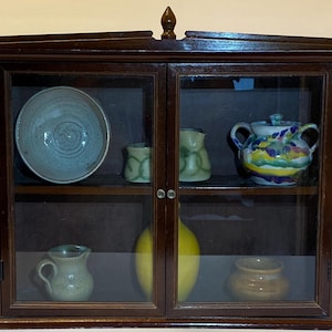 May include: A dark wood display cabinet with glass doors, showcasing various ceramic pieces. The cabinet contains a large blue-green bowl, small pitchers, a colorful sugar bowl, and a yellow sphere. The cabinet is a decorative storage solution.