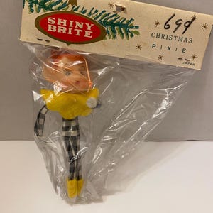 May include: A vintage Christmas pixie ornament in a plastic bag. The pixie has an orange head, yellow collar, and black and white plaid legs. The packaging reads "SHINY BRITE" and "CHRISTMAS PIXIE".