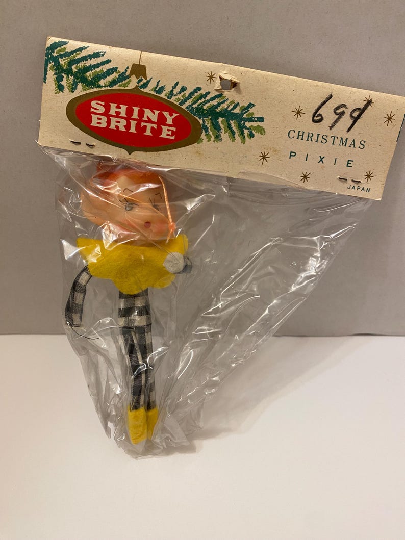 May include: A vintage Christmas pixie ornament in a plastic bag. The pixie has an orange head, yellow collar, and black and white plaid legs. The packaging reads "SHINY BRITE" and "CHRISTMAS PIXIE".