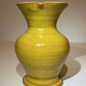 Puede incluir: Un jarrón de cerámica amarilla con un borde acampanado y un cuerpo redondeado. El jarrón tiene una base pequeña y un acabado liso y brillante. El jarrón mide aproximadamente 18 cm de alto.