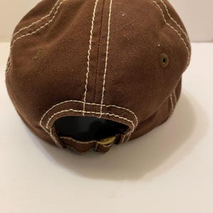 Puede incluir: Gorra de b&eacute;isbol marr&oacute;n con costuras blancas. La gorra tiene una visera curva y un bot&oacute;n en la parte superior. La parte trasera de la gorra tiene una correa ajustable con hebilla met&aacute;lica. Fabricada en tejido resistente.