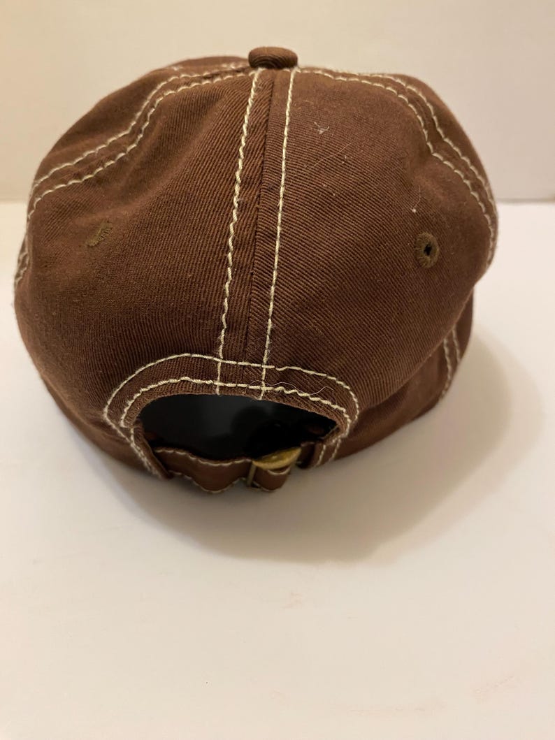Puede incluir: Gorra de b&eacute;isbol marr&oacute;n con costuras blancas. La gorra tiene una visera curva y un bot&oacute;n en la parte superior. La parte trasera de la gorra tiene una correa ajustable con hebilla met&aacute;lica. Fabricada en tejido resistente.