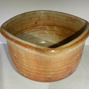 May include: A handmade, ceramic bowl with a unique, angular design. The bowl features a warm, earthy color palette with shades of tan, brown, and orange. The interior is a lighter tan, contrasting with the darker rim and exterior. The bowl is likely used for serving or display.