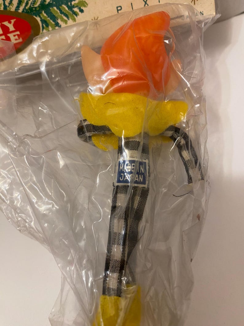 May include: A vintage pixie doll in a clear plastic package. The doll has an orange head, yellow collar, and a black and white checkered stem. The tag reads "MADE IN JAPAN".