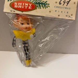 May include: A vintage Christmas pixie ornament in a clear plastic bag. The pixie has a yellow collar, black and white checkered legs, and yellow shoes. The packaging reads "SHINY BRITE" and "CHRISTMAS PIXIE".