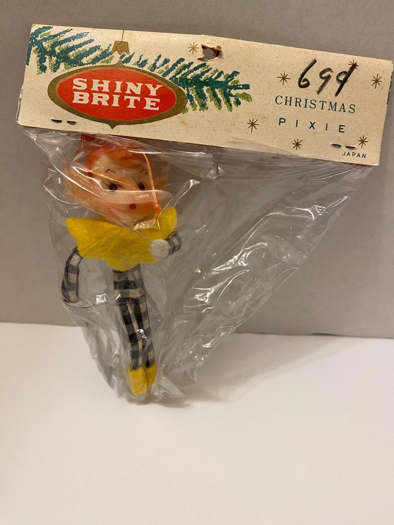 May include: A vintage Christmas pixie ornament in a clear plastic bag. The pixie has a yellow collar, black and white checkered legs, and yellow shoes. The packaging reads "SHINY BRITE" and "CHRISTMAS PIXIE".