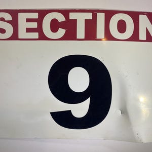 May include: A rectangular metal sign with the word "SECTION" in white letters on a red background. Below, a large black number "9" is centered on a white background. The sign has some wear and tear.