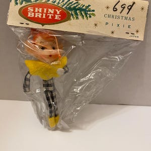 May include: A vintage Christmas pixie ornament in a plastic bag. The ornament has a yellow collar, black and white checkered pants, and yellow shoes. The packaging reads "Shiny Brite" and "Christmas Pixie."